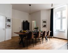 Esszimmer mit einem großen Holztisch, der von mehreren braunen Stühlen umgeben ist, in einem Raum mit Parkettboden und weißen Wänden. An den Wänden hängen gerahmte Bilder, ein großes Fenster mit weißen Vorhängen lässt Tageslicht herein, und eine schwarze Kommode steht in der Ecke.