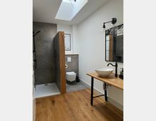 Modernes Badezimmer mit hellem Holzfußboden und großer Dachfensteröffnung. Neben einer offenen Dusche mit dunklen Fliesen befinden sich ein wandhängendes WC und ein hölzernes Waschtisch mit Aufsatzwaschbecken und Wandspiegel.