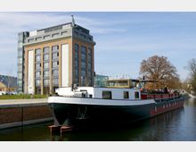 Ein modernes Backsteingebäude mit großen Fenstern steht am Ufer eines Kanals, an dem ein längliches Boot mit weißem und schwarzem Rumpf angedockt ist. Im Hintergrund sind ein kahler Baum, ein weiteres Gebäude sowie ein gepflegtes Grünflächenareal zu sehen.