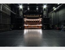 Blick von der Bühne eines Theatersaals in den Zuschauerraum mit mehreren Rängen aus Holz und beleuchteten Sitzreihen. Im Vordergrund ist die dunkle Bühne von technischen Einrichtungen und Seilzügen umgeben.