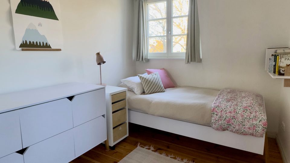Helles, minimalistisches Schlafzimmer mit weißem Einzelbett unter einem Fenster mit Vorhängen, Holzboden und hellem Teppich. An der Wand stehen eine weiße Kommode und ein kleiner Nachttisch mit Lampe; daneben hängen ein Wandbild und ein kleines Regal.