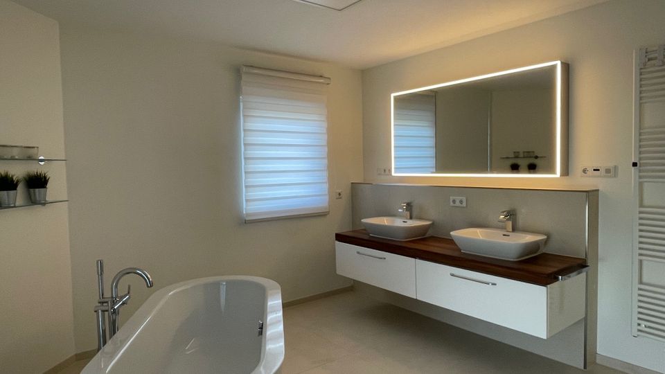 Modernes Badezimmer mit freistehender Badewanne, Doppelwaschbecken auf einer Holzplatte und beleuchtetem Wandspiegel. Das Zimmer hat helle Wände, eine mattierte Fenstergardine und einen Handtuchheizkörper rechts.