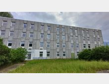 Flaches, graues Gebäude mit vier Etagen und einer Fassade aus strukturiertem Beton, unterbrochen durch rechtwinklige Fensterreihen. Im Vordergrund befindet sich eine Wiese mit hohem Gras und Sträuchern, der Himmel ist leicht bewölkt.