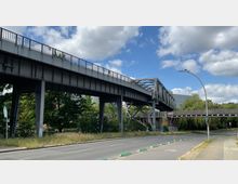 Erhöhte Stahlbrücke mit Geländern, darunter eine Straße mit Radwegmarkierungen; im Hintergrund sind Bäume, ein modernes Gebäude und ein klarer Himmel mit wenigen Wolken zu sehen.