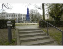 Steintreppe mit Geländer in einem Park, der zu einer offenen Fläche mit Blumenbeeten und einer blauen, pyramidenförmigen Skulptur führt; im Hintergrund stehen mehrstöckige Gebäude und kahle Bäume.