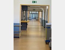 Ein langer Krankenhausflur mit gelb-braunem Boden, gelben Türrahmen und blauen Wänden. Der Flur ist mit Handläufen, großen Fenstertüren im Hintergrund und Notausgangsschildern ausgestattet; rechts stehen Reinigungswagen.