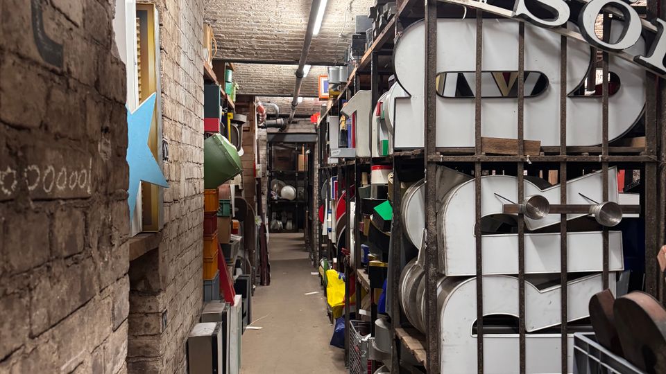 Ein schmaler Lagerraum mit Backsteinwänden und gewölbter Decke. Regale voller großer Buchstaben, bunter Objekte und Kisten säumen den Gang, der von Neonröhren beleuchtet wird.