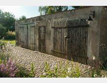 Steingepflasterter Hof mit einer einfachen rustikalen Scheune aus rohem Putz und Holztüren, umgeben von Gartenpflanzen und grüner Vegetation im Hintergrund. Ein Wandlicht hängt an der rechten Seite des Gebäudes.