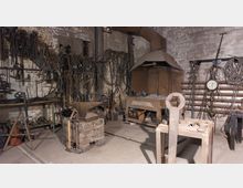 Traditionelle Schmiede mit Amboss, Feuerstelle und zahlreichen Werkzeugen an den Wänden. Der Raum hat unverputzte Backsteinwände und ist mit Arbeitsutensilien wie Hämmern, Ketten und Schraubenschlüsseln ausgestattet.