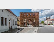 Backstein-Stadttor in einer historischen Altstadt, flankiert von Gebäuden mit heller Fassade und roten Dächern; im Hintergrund eine Kirche mit zwei Türmen. Die Straße führt durch das Tor, umgeben von gepflasterten Gehwegen und Straßenlaternen.