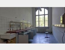 Altes Labor mit einer langen Arbeitsfläche, weißen Fliesenwänden und mehreren Wasseranschlüssen. Im Hintergrund befindet sich ein großes Fenster mit Blick auf Bäume, darunter ein Heizkörper; offene Schränke und ein einfacher Hocker ergänzen die Einrichtung.