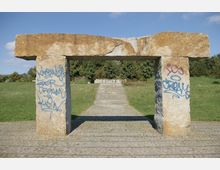 Steinernes Tor aus großen, grob behauenen Steinblöcken mit Graffiti, davor ein gepflasterter Weg und dahinter eine Treppe, die zu einer Grünfläche mit Bäumen führt. Der Himmel ist klar und blau.