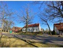 Straßenansicht in einem Dorf, mit einer gepflasterten Straße, grasbewachsenen Flächen und mehreren Bäumen ohne Blätter. Im Hintergrund stehen Wohnhäuser mit roten Ziegeldächern, darunter ein weißes Gebäude im traditionellen Baustil.