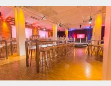 Veranstaltungsraum mit modernen hohen Tischen und Barhockern, umgeben von Backsteinwänden und beleuchtetem Deckenraster. Im Hintergrund befindet sich eine kleine Bühne mit Bildschirmen und bunten LED-Lichtern.