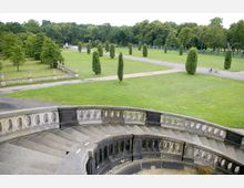 Weitläufige Parkanlage mit gepflegtem Rasen, vereinzelten hohen Bäumen und symmetrisch angelegten Wegen. Im Vordergrund befindet sich eine steinerne, halbkreisförmige Treppe mit Geländer, während im Hintergrund eine Allee und dichte Baumreihen zu sehen sind.