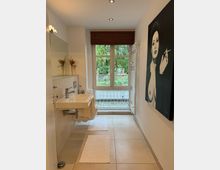 Modernes Badezimmer mit weißer Ausstattung, rechteckigem Waschbecken und großem Fenster mit Blick auf einen Garten. An der rechten Wand hängt ein großes Schwarz-Weiß-Gemälde, und der Boden ist mit beigefarbenen Fliesen ausgelegt.