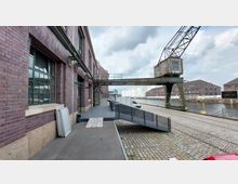 Industriegelände mit einer Backsteinfassade links, versehen mit großen Fenstern. Im Hintergrund ein alter Kran über Bahngleisen, angrenzend an einen Wasserkanal mit Speichergebäuden und weißen Tanks auf der rechten Seite.