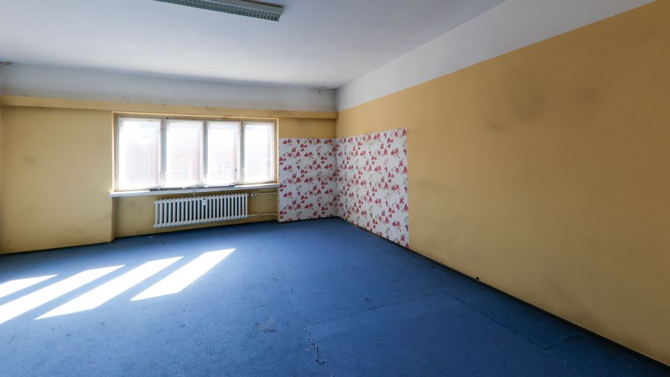 Leerer Innenraum mit blauem Boden, beigefarbenen Wänden und großer Fensterfront mit Heizkörper darunter. An einer Ecke befindet sich ein Abschnitt mit rot gemusterter Tapete, an der Decke hängt eine lange Leuchtstofflampe.