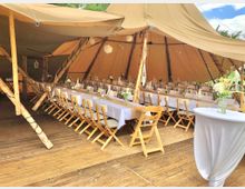 Großes Festzelt mit beigefarbener Zeltplane und einer Holzkonstruktion, innen mit langen, weiß gedeckten Tischen und Holzstühlen arrangiert. Auf den Tischen stehen Blumenarrangements, und der Boden besteht aus Holzplanken.