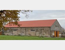 Ein langgestrecktes Gebäude mit Ziegeldach und unverputzten Steinwänden, vermutlich ein landwirtschaftlicher Schuppen oder eine Lagerhalle. Im Vordergrund eine Wiese, im Hintergrund ein bewölkter Himmel, links ein Baum mit herbstlichem Laub.
