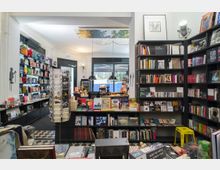 Ein kleiner Buchladen mit bodenständigen, dunklen Regalen, die mit Büchern, Postkarten und Geschenkartikeln gefüllt sind. Im Hintergrund beleuchten Hängeleuchten den Kassenbereich, während durch große Fenster Tageslicht einfällt.