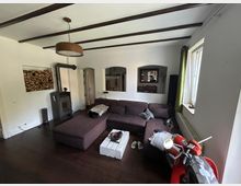 Wohnzimmer mit dunklem Holzboden, einer großen dunkelbraunen Eckcouch, einem weißen Couchtisch aus Paletten und einem Holzofen mit gestapeltem Brennholz in der Wandnische. Auf der rechten Seite ein Fenster mit grünem Vorhang und im Vordergrund ein roter Motorroller.