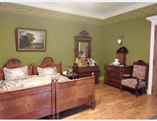 Historisches Schlafzimmer mit Gründerzeitmöbeln: Zwei hölzerne Einzelbetten mit geschnitzten Kopf- und Fußteilen, ein passender Spiegelkommode, dekorative Wandbordüre auf olivgrüner Wand, Parkettboden und antike Accessoires wie ein Polsterstuhl und Waschutensilien.