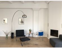 Wohnzimmer mit minimalistisch-moderner Einrichtung, bestehend aus einem schwarzen Ledersessel mit passendem Fußhocker, einem Drahtsessel mit blauem Kissen, einem kleinen Holztisch mit Pflanze und einer geschwungenen Stehlampe aus Metall. Die Wände sind weiß, der Boden ist aus hellem Holz, und rechts befindet sich ein in die Wand integrierter Kamin.