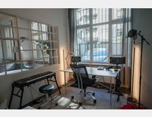 Ein Arbeitszimmer mit einem Schreibtisch, einem Bürostuhl, einem Mikrofon und Lautsprechern. Im Raum befinden sich zudem ein digitales Keyboard mit Hocker und eine große Fensterfront mit Gitter, durch die ein Innenhof zu sehen ist.