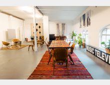 Großzügiger, modern eingerichteter Raum mit weiß gestrichenen Wänden, rustikalem Holztisch, passenden Stühlen und buntem Teppich. Im Hintergrund befinden sich gemütliche Sitzbereiche mit Teppichen, Pflanzen und Wanddekorationen, darunter Schwarz-Weiß-Fotografien und geometrische Musterteppiche.