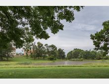 Weitläufige Parklandschaft mit einem kleinen Teich in der Mitte, umgeben von grünen Wiesen und Bäumen; im Hintergrund sind helle Gebäude mit roten Dächern zu sehen. Der Himmel ist wolkenbedeckt, und überhängende Äste rahmen das Bild ein.