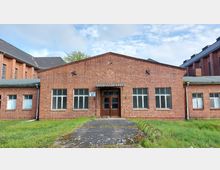 Backstein-Gebäude mit flachem Dach, großflächigen Fenstern und einem zentralen Eingang mit Doppeltür, beschriftet als "Gebäude 31." Umgeben von einer Rasenfläche, mit weiteren angrenzenden Backsteinbauten im Hintergrund.