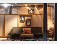 Gemütlicher Innenraum mit Backsteinwand, in die mehrere Schwarz-Weiß-Fotografien integriert sind. Der Raum ist mit einer schlichten Holzbank, einem niedrigen Holztisch, Hockern und einem Ledersessel eingerichtet, während sichtbare Rohre und Beleuchtung eine industrielle Atmosphäre schaffen.