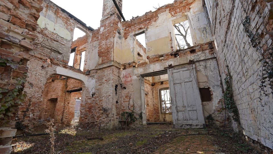 Innenansicht einer verfallenen Backsteinruine ohne Dach, mit freiliegenden Mauerresten, hohen Fensteröffnungen und einem teils erhaltenen Rundbogen. Im Vordergrund liegen überwucherte Ziegelböden und eine schief stehende Tür im offenen Durchgang.