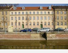 Historisches Gebäude mit pastellgelber und pfirsichfarbener Fassade sowie grünen Türen, flankiert von zwei ähnlichen Gebäuden. Im Vordergrund sind parkende Autos und ein Wasserkanal mit einer kleinen Treppe zu sehen.