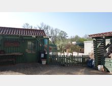 Grüner Gartenbereich mit einer kleinen Holzhütte mit Ziegeldach, Holzzaun und gepflastertem Boden. Im Hintergrund sind blühende Bäume, ein kleiner Teich und weitere Gebäude sichtbar.