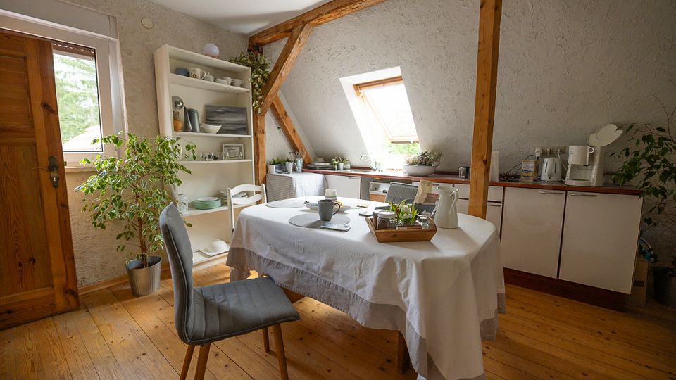 Gemütlicher Küchen- und Essbereich im Dachgeschoss mit sichtbaren Holzbalken, Holzfußboden und weiß verputzten Wänden. Ein runder Tisch mit Tischdecke steht vor einer Küchenzeile unter einem Dachfenster; daneben ein offenes Regal, Zimmerpflanzen und ein seitliches Fenster.