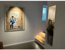 Flur mit moderner, heller Innenausstattung, das rechteckige Kunstwerk an der Wand zeigt einen Street-Art-Stil mit der Aufschrift „Banksy is a girl“. Rechts befindet sich eine offene Treppe mit beleuchteten Stufen, daneben eine dekorative Nische mit einem hohen Pflanzenarrangement.