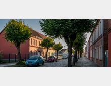 Straßenszene mit Kopfsteinpflaster, gesäumt von Bäumen und zweigeschossigen Ziegelhäusern. Am Straßenrand stehen parkende Autos, und im Hintergrund sind weitere Gebäude sichtbar.