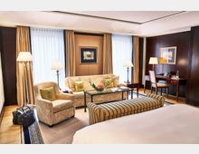 Elegantes Hotelzimmer mit hellen Polstermöbeln, einem Glastisch mit Blumendekoration und einer Arbeitsnische mit Schreibtisch und Stuhl; bodenlange Vorhänge vor großen Fenstern und dunkelbraune Holzverkleidung an den Wänden. Im Vordergrund ein Bett und eine gestreifte Sitzbank.