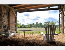 Innenansicht einer offenen Scheune mit Blick auf eine Wiesenlandschaft und Bäume im Hintergrund. Im Vordergrund stehen zwei weiße Holzstühle und ein kleiner Tisch auf einem Boden aus Stroh.