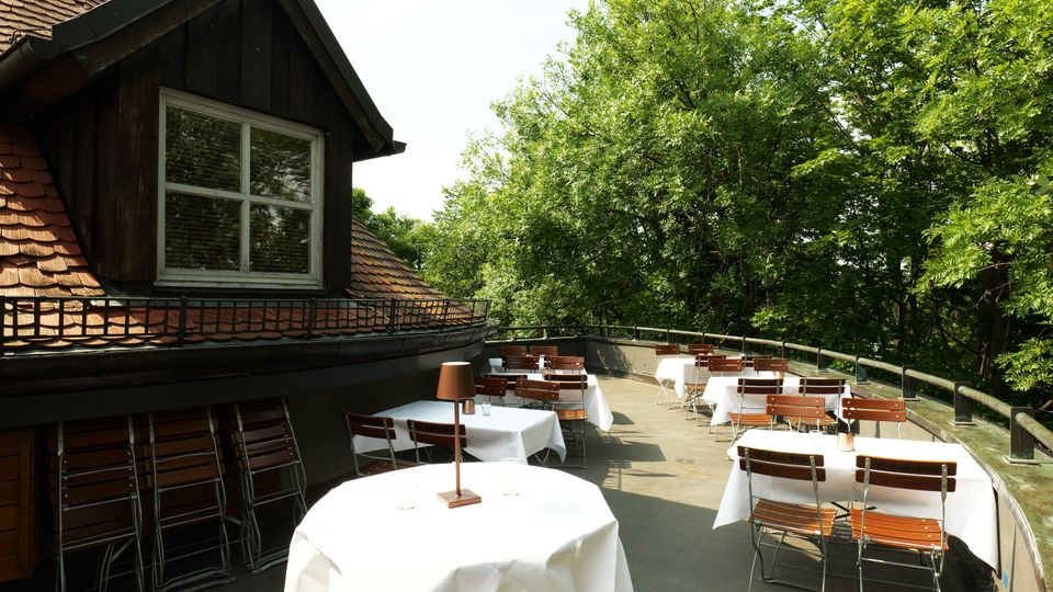 Dachterrasse eines Gebäudes mit Schindeldach und Gaube, ausgestattet mit Tischen mit weißen Tischdecken sowie Holzstühlen. Entlang der Brüstung verläuft ein Geländer; die Terrasse ist von dichtem, grünem Baumbestand umgeben.
