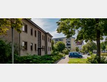 Mehrfamilienhäuser in einem Wohngebiet mit einer Grünfläche und Gehweg im Vordergrund, Bäumen sowie parkenden Autos; im Hintergrund ein modernes Wohngebäude mit Balkonen.