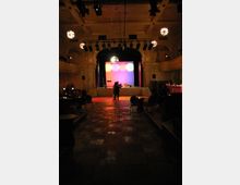 Ein eleganter Veranstaltungssaal mit hohen Decken und einer beleuchteten Bühne im Hintergrund, die bunte Kreiseffekte zeigt. Im Vordergrund tanzt ein Paar auf einer dunklen Parkettfläche, umgeben von runden Tischen mit Kerzenlicht.