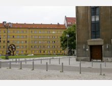 Ein gepflasterter Vorplatz mit Metallpfosten und Straßenlaternen, im Hintergrund ein gelbes Wohngebäude mit mehreren Stockwerken und einem roten Dach. Rechts im Bild ist ein Teil eines großen Gebäudes mit einer Holztür und einem hohen Fenster sichtbar.