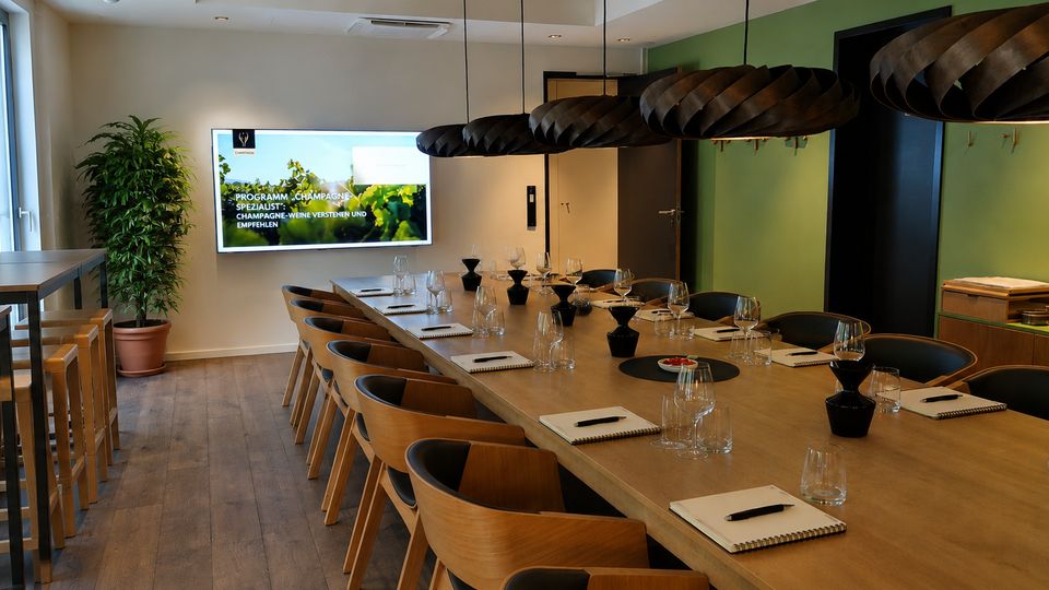 Moderner Seminar- und Verkostungsraum mit langem Holztisch, Stühlen und eingedeckten Gläsern sowie Notizblöcken. An der Stirnwand hängt ein Bildschirm, daneben steht eine große Topfpflanze; über dem Tisch hängen mehrere dunkle, runde Pendelleuchten, links sind hohe Fenster und Barhocker an einer Theke.