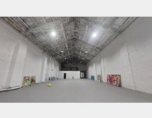 Großer, leerer Industrie-Raum mit weiß gestrichenen Wänden und offenem Dachstuhl aus Holz und Metall. Entlang der Wände sind vereinzelt bunte Gemälde und Gegenstände abgestellt.