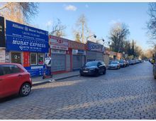 Eine gepflasterte Straße mit parkenden Autos rechts und links; links befinden sich kleine Ladenlokale mit Werbeschildern, darunter ein Versicherungsbüro und ein Zulassungsdienst. Im Vordergrund steht ein Mann mit einem Elektroroller vor einem der Geschäfte. Im Hintergrund sind Bäume zu sehen.