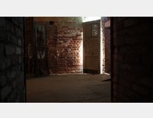 Ein dunkler Raum mit freiliegenden Ziegelwänden, einem alten Metallgitter an der linken Wand und einer rustikalen Holztür mit einem kleinen perforierten Fenster, durch das Licht einfällt. Der Boden ist aus Beton, und der Raum wirkt vernachlässigt oder verlassen.