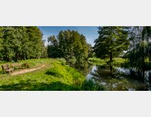 Ein natürlicher Park mit einem schmalen Weg, der an einer Reihe von Holzbänken entlangführt, daneben ein ruhiger Fluss mit Bäumen am Ufer. Die Umgebung ist geprägt von dichter Vegetation, darunter hohe Bäume und grasbewachsene Flächen.
